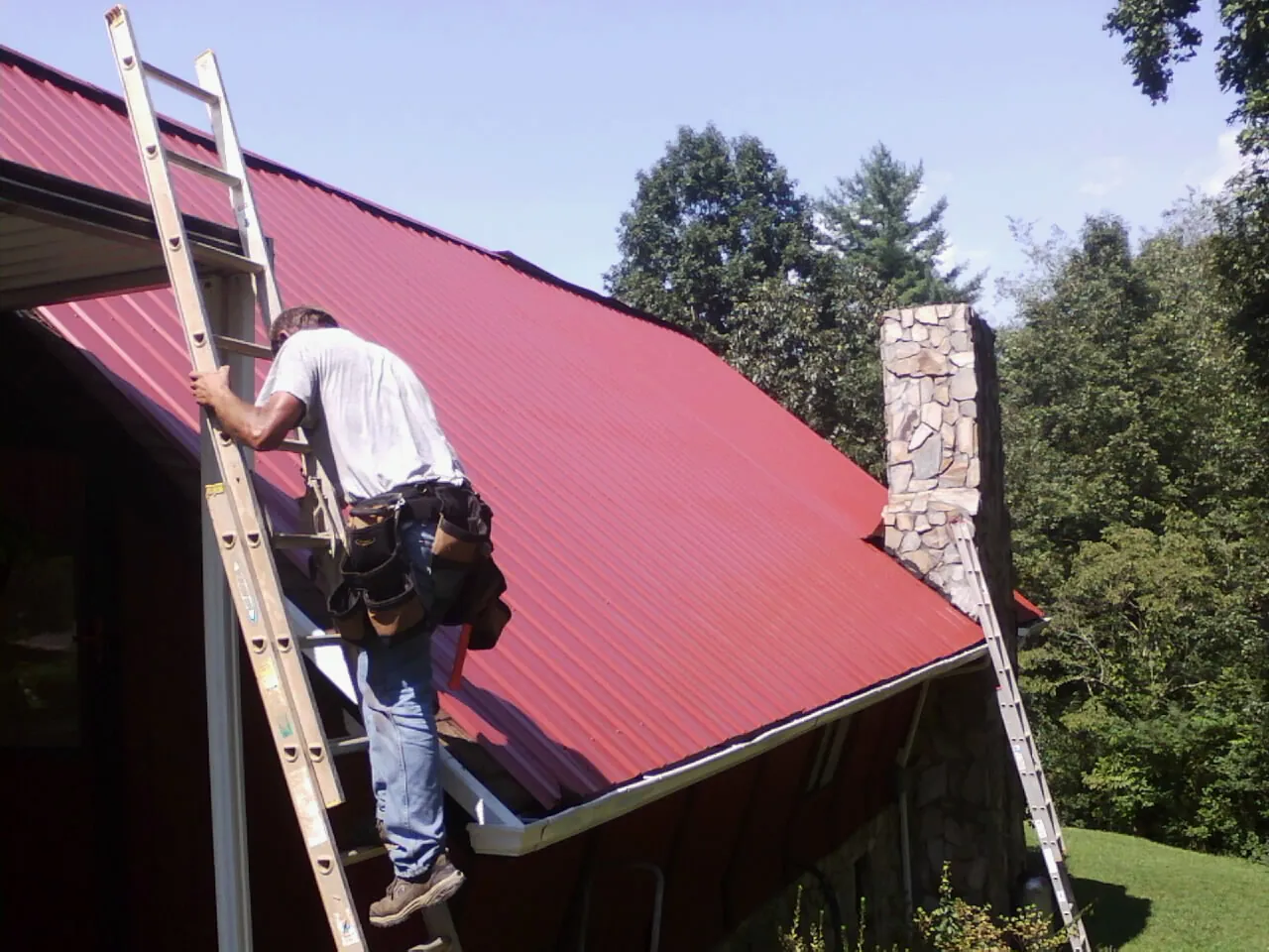 Roofer on a ladder beside a red metal roof and stone chimney — Soffit & Fascia Repair by Roofing Brothers in North Arlington